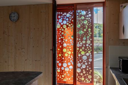 Architektonisches Schiebepaneel in Orange mit kunstvoller, kreisförmiger Perforation vor einer vertikalen Holzfassade. Detailaufnahme der Materialkombination.