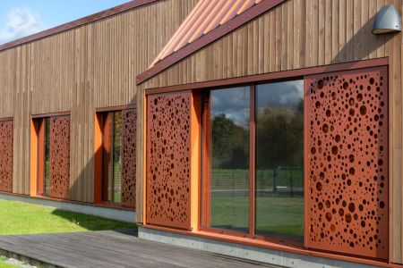 Orangefarbener Schiebeladen aus Aluminium mit Stanzmuster vor einer Holzfassade.