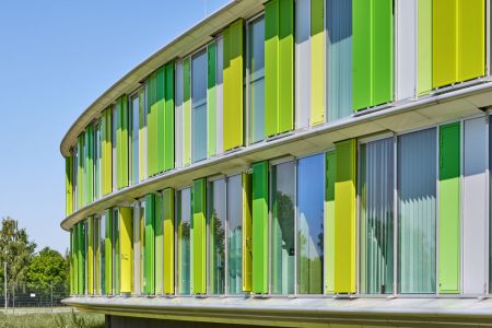 Gebogene Gebäude-Fassade mit grünen Faltschiebeläden und Schiebeläden aus perforiertem Aluminium als Sonnenschutz.
